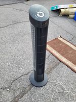 Black tower stand up fan photographed on outdoor asphalt surface, showing the entire fan body including controls, vents and power cord on ground.