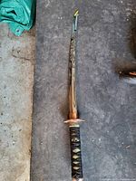 Top-down view of a Japanese katana sword laid flat on the ground. Blade has a dragon design engraved, handle is traditionally wrapped in black with gold diamond patterns, copper-colored guard visible.