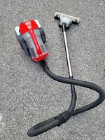Red and gray Dirt Devil Power Reach vacuum cleaner on pavement showing overall unit with dust container, hose, and floor brush attachment.