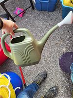 The photo shows a green plastic watering can with a white spout held by a hand above the ground. Other items like a blue storage bin and a yellow rope are partially visible in the background.