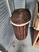 Tall brown wicker laundry basket with hinged lid showing side view in storage area