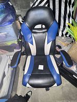 Front-facing view of a black, blue, and white gaming-style desk chair, showing seat, backrest with a ventilation/design hole near the top, and padded armrests.