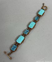 Full length view of gold-tone bracelet with alternating carved turquoise stones and blue faceted stones on gray background