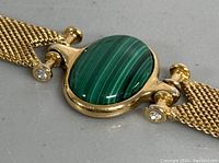 Close-up of large oval malachite stone set in gold-tone metal with mesh band and rhinestone accents.