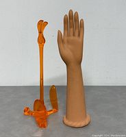 Front view of vintage mannequin hand arm next to amber-orange plastic stand on gray surface against white background
