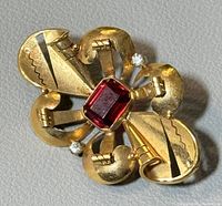 Front top view of yellow gold brooch showing four-lobed scroll design, emerald-cut red center gemstone, two round accent stones