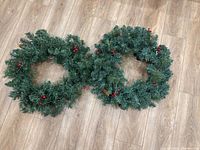 Photo showing two green artificial Christmas wreaths placed on a wooden floor, featuring red berry decorations and string lights.