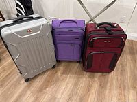 Photo of three suitcases including a large silver hard-shell one, a medium purple soft-sided one, and a smaller maroon soft-sided one, placed on a hardwood floor.