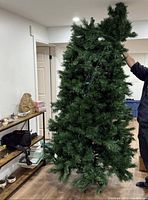 Partial view of the artificial Christmas tree unassembled indoors, held upright by a person, showing dense green branches and prelit integrated lights wiring.