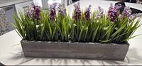 Photo of a rectangular wooden planter box filled with artificial lavender flowers and green foliage placed on a white table near a window.