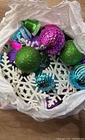 Top view of assorted Christmas ornaments in white plastic bag including green, purple, blue glitter spheres, and white snowflakes.