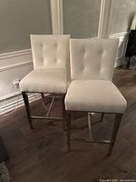 Pair of two white upholstered bar stools with button tufting on the backs and seats, metallic legs with footrest bars, standing on dark wood floor.