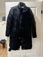 Front view of black Calvin Klein long padded ski jacket with zipper and snap button closure, side pockets, hanging on white door.