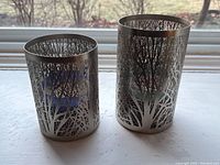 Two silver cylindrical candle holders with intricate cutout designs of tree branches on a window sill.