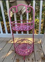 Front view of a two-tier cast iron plant stand painted pink but with chipped paint and some rust, placed on wooden deck outdoors.