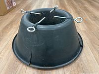 Black plastic tapered Christmas tree stand with four metal eye bolts for securing tree trunk, showing some dust and wear.