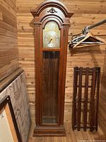 Full frontal view of the grandfather clock showing the wooden arched top, brass clock face with black hands, and glass pendulum door. Clock is missing weights below the pendulum.