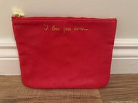 Red zipper pouch with gold text 'I love you more...' against white wall on wooden floor