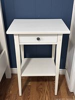 White IKEA night stand with one drawer and lower shelf, standing on wooden floor against dark blue wall.
