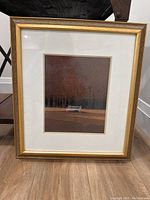 Photo of gold framed print depicting a white solo bench with trees in the background, with visible white matting and gold frame.
