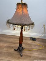 Full view of the table lamp showing its beige fabric shade with fringes, wooden body, and metal tripod base on a wooden floor against a white wall.