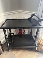 Photo of a black painted vintage wooden tea cart with two shelves, turned wood legs, a bottle holder on bottom shelf, and wheels for mobility.