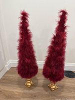 Photo showing two tall red feather Christmas trees with gold bases against white wall on wood floor.