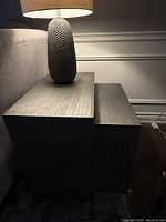 Side view of two nested greyish silver end tables with textured finish and lamp on top.