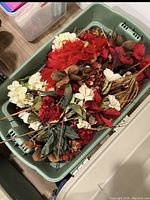 Green plastic bin filled with various artificial Christmas flowers including poinsettias, white flowers, brown seed pods, and red berries.