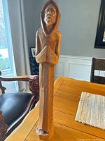 Front view of a hand carved wooden statue showing a hooded figure with folded hands in prayer.