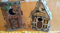 Two tin bird houses on a table: one outhouse style with crescent moon cutout and artificial ivy, the other a house with 'Bed & Breakfast' sign and shingled roof.