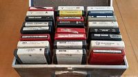 Photo showing 24 eight-track tapes in a gray storage box, labels visible with artist names and condition markings.