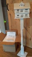 Full view of the white Cape Cod birdhouse mounted on the pedestal stand, showing front facade and packaging box with instructions.