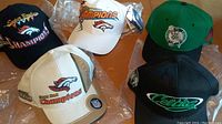 Front view of two Denver Broncos Super Bowl Champions hats in white and beige, and two Boston Celtics hats in black and green.