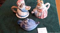 Overview photo showing three collectible ceramic teapots placed on a green fabric surface: two cat-shaped teapots wearing dresses and bonnets, one pink and one blue, and one blue floral teapot with a bird-shaped lid.