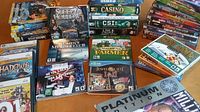 Wide view of multiple PC game boxes stacked and spread out on a table showing various titles and genres including casino, mystery, puzzle, farming, and more.