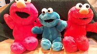 Photo showing two red Tickle Me Elmo plush toys and one blue Tickle Me Cookie Monster plush toy seated on a wooden surface in front of a green fabric background, capturing front view of toys.