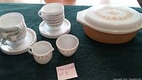 Full lot photo showing six stacked cups, matching saucers, creamer jug, sugar bowl, and covered casserole on a dark green cloth.