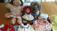 Photo showing three vintage dolls including Cabbage Patch, Raggedy Ann, and Wrinkles, with clothing spread on table.