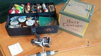 Wide shot showing tackle box filled with vintage lures, bait box, and jigging rod with reel placed on a wooden surface