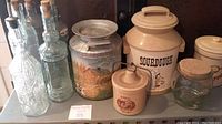 Photo showing 3 embossed glass bottles, metal tin with landscape, large sourdough ceramic crock, small marjoram ceramic jar, corked clear glass jar, and cream crock