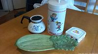 Overall view of all four vintage kitchenware items on a wooden table: Kellogg's cookie jar, Corning Ware pot, Wonder Bread container, and celery dish.