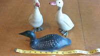 Photo showing three wooden duck figurines; two white ducks with orange beaks and metal feet, one black loon with white spots. Tape measure for scale shows about 11 inches for the loon.