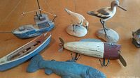 Wide shot showing all 6 wooden sea shore decor items arranged on wood floor: two small model boats, two carved shorebirds, wooden lure, and carved fish sculpture.