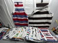 Two striped tank tops hung showing one white with black and brown stripes, and one white with red, blue, and maroon stripes. Multiple tank tops packaged below.