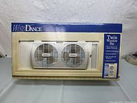 Full front view of boxed WindDance Twin Window Fan showing front of packaging and the fan unit inside with dual fan blades visible.