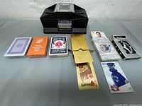 Photo showing five decks of playing cards and one black electric card shuffler on a gray surface.