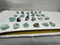 Lot of 24 raw amazonite stones displayed on a white cloth with a measuring tape showing size scale. The stones vary in size and shape, showcasing their natural rough form and light blue-green Amazonite color.