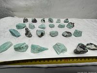 24 raw Amazonite stones laid out on a white surface with a yellow measuring tape below showing approximate scale.