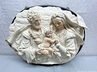 Front view of the religious wall mount figure showing detailed sculpted relief of the Holy Family in cream color with gold accents, mounted on oval plaque.